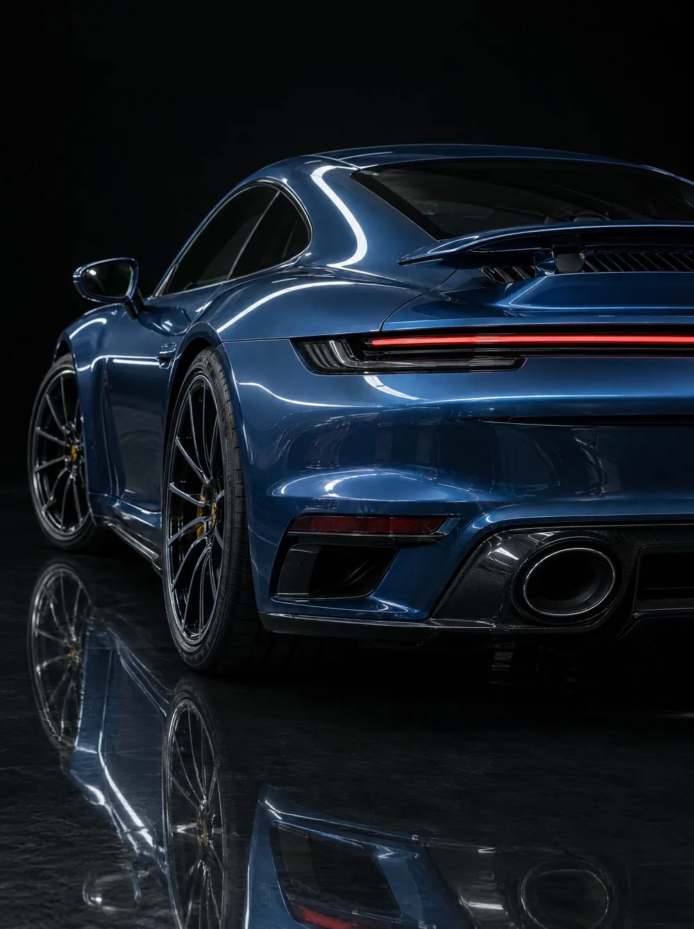 The glossy rear three-quarter of a deep blue Porsche 911 Turbo S, a curved highlight running the length of its roofline and its reflection in the dark floor.