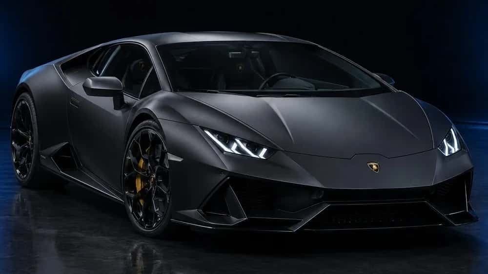 A satin black Lamborghini Huracán across a wide frame, the light from the panel overhead spread into a soft sheen over its matte body instead of a sharp reflection.