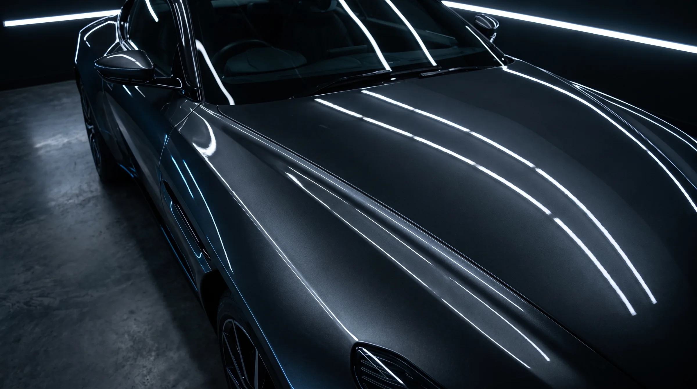 A dark gunmetal grey sports car's long hood and front fender seen from above the front corner, the paint deep and unmarked, the studio's white light bars reflecting in long unbroken lines.