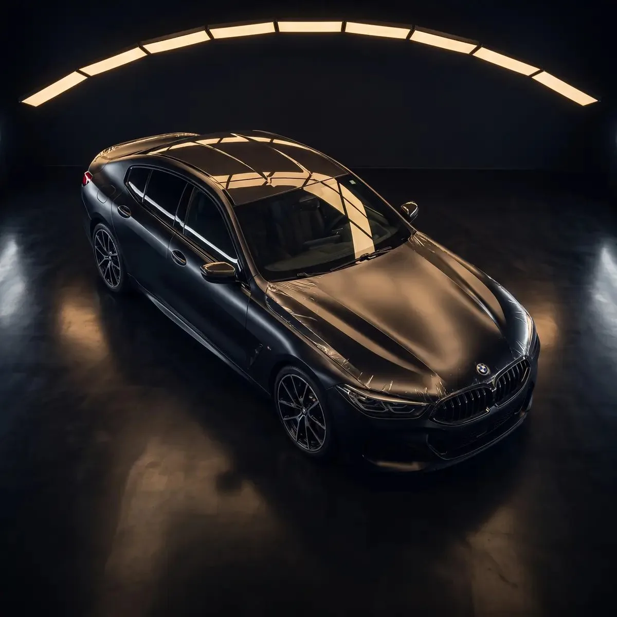 A dark BMW 8 Series seen from above with a sheet of clear film laid across its hood and roof, under a long arc of warm ceiling light panels.