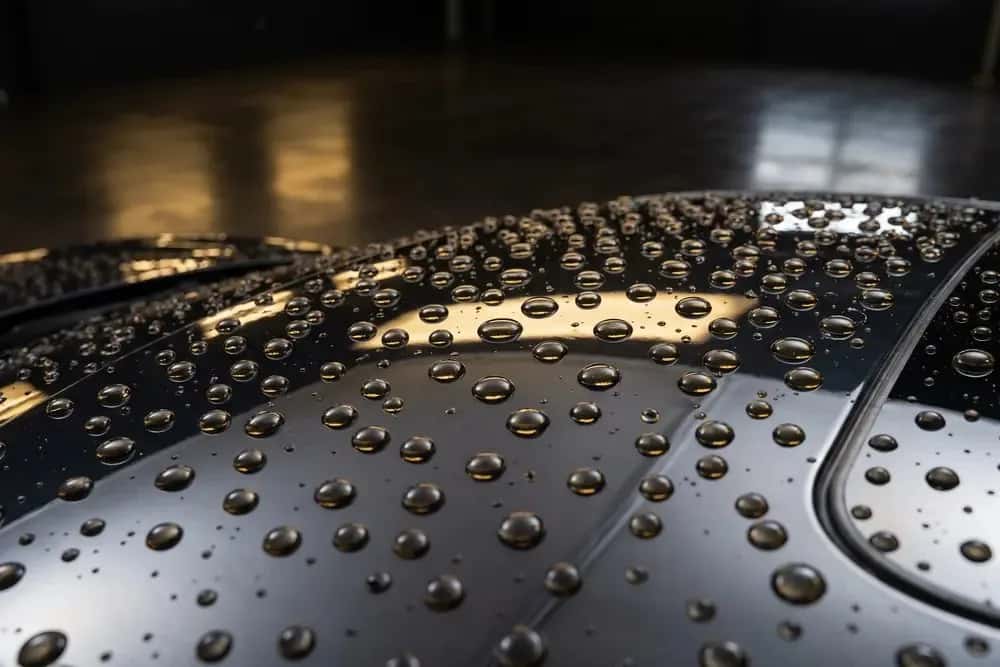 Close-up of water beads standing on the black rear deck of a car, warm ceiling lights mirrored in the wet paint behind them.