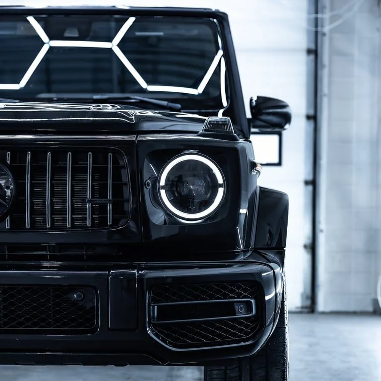 The front of a black Mercedes G-Class, its round headlight ring lit and hexagonal light panels reflected across the windshield.