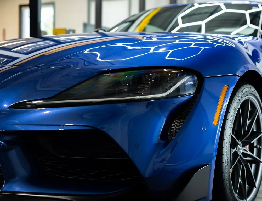 The front corner of a blue Toyota GR Supra, hexagonal ceiling lights reflected across its hood and fender.